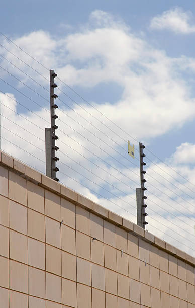 Electric fence at the top of a wall, against a cloudy blue sky.